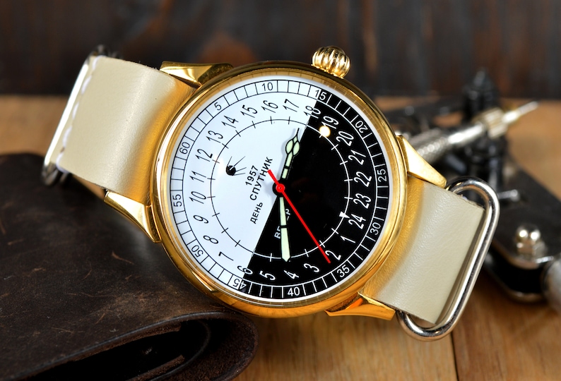 May include: A gold-tone wristwatch with a white and black face. The face has a 24-hour clock with the text "День Спутник 1957" (Day Satellite 1957). The watch has a white leather band.