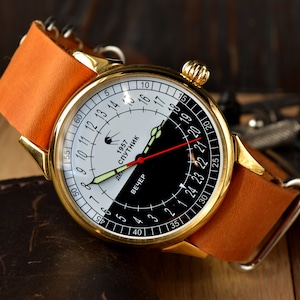 May include: A gold-tone wristwatch with a black and white face. The face has a 24-hour clock with the words "1957 Sputnik" and "Вечер" (Evening) printed on it.