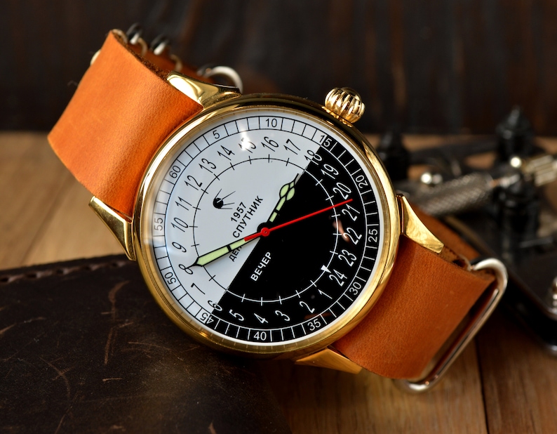 May include: A gold-tone wristwatch with a black and white face. The face has a 24-hour clock with the words "1957 Sputnik" and "Вечер" (Evening) printed on it.