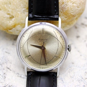 Może przedstawiać: Srebrny zegarek na rękę z czarnym skórzanym paskiem. Tarcza zegarka jest srebrna z czarnymi oznaczeniami i tekstem "Raketa 21 JEWELS MADE IN USSR".