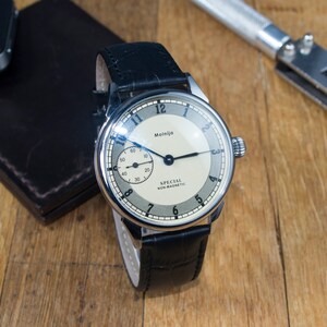 Op de afbeelding: Een zilveren polshorloge met een witte wijzerplaat en zwarte wijzers. Het horloge heeft een zwarte leren band en staat "Molnija" en "Special Non-Magnetic" op de wijzerplaat.
