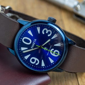 Peut inclure: Une montre-bracelet à cadran bleu avec des chiffres et des aiguilles blancs, et le mot "PAKETA" sur le cadran. La montre a un bracelet en cuir marron et un fermoir argenté. La montre est sur une surface marron.