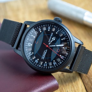 May include: A black wristwatch with a mesh band. The watch face is black with white numerals and a blue and red design. The watch hands are red and white. The watch is on a burgundy surface.