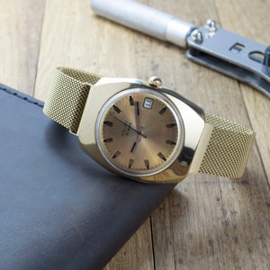May include: A gold-toned vintage wristwatch with a mesh band rests on a dark gray surface. The watch has a gold face with black markers and a date window. A watch repair tool is in the background, suggesting watchmaking or repair.