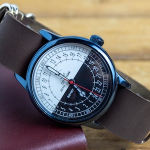 Puede incluir: Un reloj de pulsera con caja de metal azul oscuro y correa de cuero marrón. La esfera está dividida diagonalmente en secciones blanca y negra con una escala de 24 horas, un segundero rojo y texto en cirílico que incluye la palabra Sputnik.