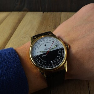 May include: A gold-tone wristwatch with a black leather band. The watch face is white and black with a red hand and the text "1957 СПУТНИК" and "ВЕЧЕР".