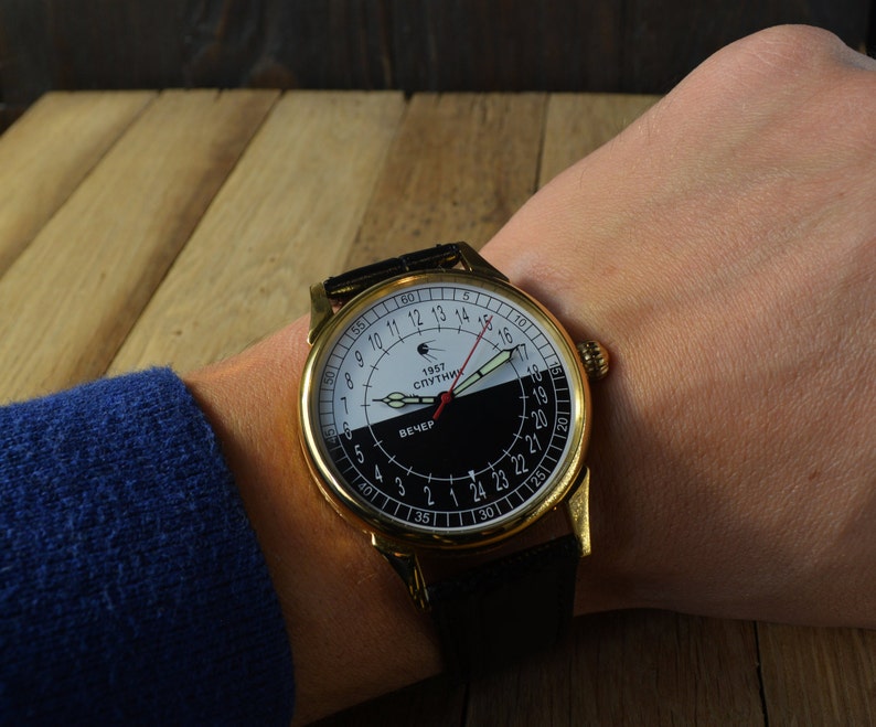 May include: A gold-tone wristwatch with a black leather band. The watch face is white and black with a red hand and the text "1957 СПУТНИК" and "ВЕЧЕР".