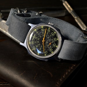 May include: A silver wristwatch with a black face and a gray leather band. The watch has a second hand and a small dial for the seconds. The watch is on a brown leather surface.