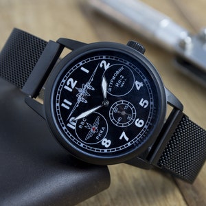 Puede incluir: Un reloj de pulsera negro con una correa de malla negra. La esfera del reloj tiene un fondo negro con marcas blancas y una silueta de un avión. El texto en la esfera del reloj dice "ШТУРМОВИК Ил-2 1941 г." y "BBC РККА".
