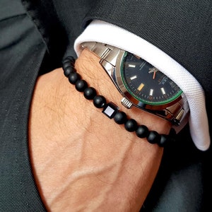 Pode incluir: Um bracelete de contas pretas com um fecho quadrado prateado no pulso de uma pessoa. O bracelete é usado com um relógio prateado com mostrador preto e detalhes verdes.