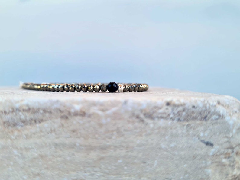 Pode incluir: Uma pulseira de contas com pequenas contas de pirita dourada e uma &uacute;nica conta de &ocirc;nix preta. A pulseira est&aacute; sobre uma superf&iacute;cie de pedra clara, com um fundo azul suave. As contas t&ecirc;m cerca de 0,3 cm de di&acirc;metro.