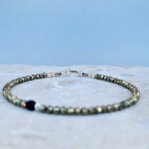 Pode incluir: Uma pulseira de contas de pirita com fecho prateado e uma &uacute;nica conta azul escura. A pulseira &eacute; feita de pequenas contas facetadas em tons de ouro e cinza. O fecho &eacute; prateado.