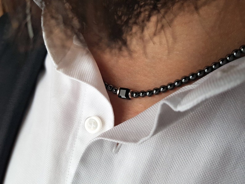 May include: A black beaded necklace with a black rectangular pendant. The necklace is worn by a person with a white collared shirt.