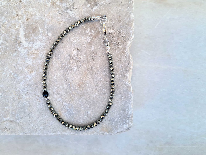 Pode incluir: Uma pulseira de contas com pequenas contas cinza met&aacute;licas facetadas. Uma &uacute;nica conta preta est&aacute; incorporada no design. A pulseira tem um fecho prateado.