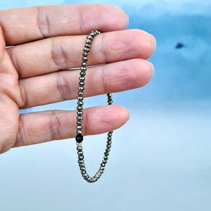 Pode incluir: Uma pulseira delicada com pequenas contas de pirita met&aacute;licas enfiadas. Uma &uacute;nica conta redonda, preta, &eacute; incorporada no design. A pulseira &eacute; segurada contra um fundo azul claro.