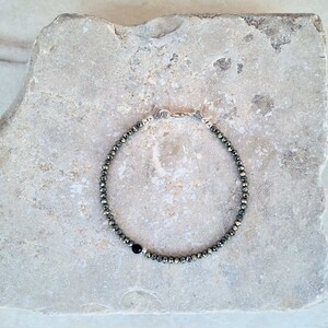 Pode incluir: Uma pulseira de contas met&aacute;licas redondas e brilhantes, de cor escura e iridescente. Uma &uacute;nica conta preta est&aacute; inserida na pulseira. A pulseira &eacute; fechada com um fecho prateado.