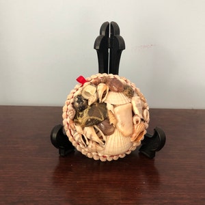 May include: A round ornament decorated with various seashells in shades of white, beige, and brown. A small red ribbon is attached to the top. The ornament is displayed on a black stand, suitable for coastal or nautical decor.