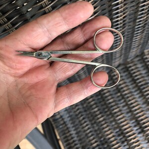May include: Small, silver-colored scissors with rounded finger holes. The blades are pointed and slightly curved. The scissors have text on the body, including the word "Miller". The scissors are designed for precision cutting.