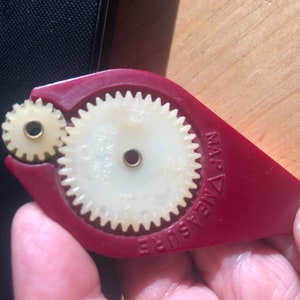 May include: A red and cream-colored map measure tool. The tool has two interlocking gears, one large and one small, with the words "MAP MEASURE" printed on the red handle. The gears are cream-colored and have a small hole in the center.