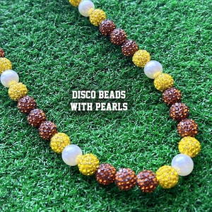 May include: A necklace made of brown, yellow and white beads. The beads are interspersed with white pearls. The necklace is on a green grassy surface. The text "DISCO BEADS WITH PEARLS" is printed on the image.