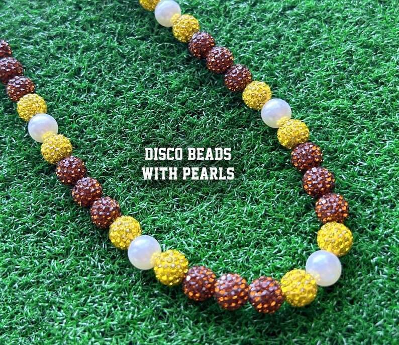 May include: A necklace made of brown, yellow and white beads. The beads are interspersed with white pearls. The necklace is on a green grassy surface. The text "DISCO BEADS WITH PEARLS" is printed on the image.