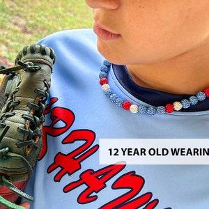 May include: A young person wearing a blue shirt with red lettering and a red, white and blue beaded necklace. The necklace is 18 inches long. The person is holding a green and brown baseball glove. "12 YEAR OLD WEARING 18" is written in white text on the shirt.