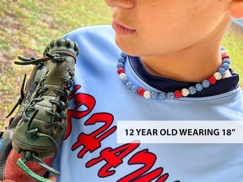May include: A young person wearing a blue shirt with red lettering and a red, white and blue beaded necklace. The necklace is 18 inches long. The person is holding a green and brown baseball glove. "12 YEAR OLD WEARING 18" is written in white text on the shirt.