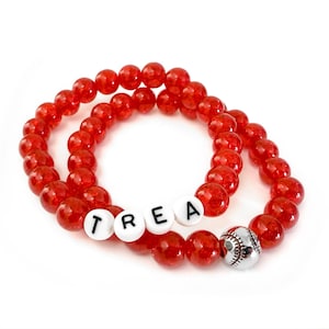 May include: Two red beaded bracelets. One bracelet has the word "TREA" spelled out in white letter beads. The other bracelet has a silver baseball charm. The bracelets are on a white background.
