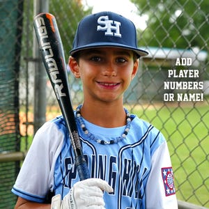 May include: A young baseball player wearing a blue and white baseball uniform with a blue hat that says "SH" on it. He is holding a black and silver baseball bat with the word "RADLIGS" on it. The player is standing in front of a chain link fence. The text "ADD PLAYER NUMBERS OR NAME!" is visible in the background.