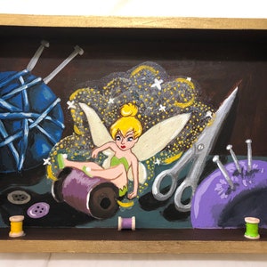 May include: A rectangular wooden box with a gold-painted frame containing a painting of a fairy with blonde hair and green clothing sitting on a purple spool of thread, surrounded by blue yarn, silver scissors, a purple pincushion, and buttons.