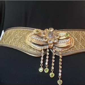 May include: A gold-tone belt with a large, ornate buckle featuring a cluster of clear and green gemstones. The buckle is attached to a brown leather strap with a crocodile-embossed pattern.