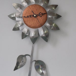 May include: A metal sunflower clock with a copper-coloured face. The clock has black hands and a silver stem with two leaves.