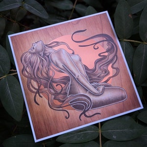 May include: Square art print featuring a nude figure with long flowing hair, rendered in shades of brown and tan. The artwork is set against a wood-grain background with a peach-colored circular element. The print has a white border.
