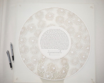 May include: A white ketubah with a circular design of delicate floral patterns. The ketubah is written in Hebrew and includes the names of the bride and groom. Two silver pens are placed on the white surface.
