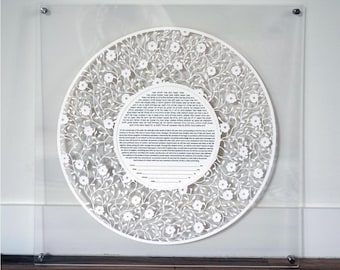 以下が含まれることがあります： 透明なアクリル製の正方形フレームに収められた、白い切り絵の円形ケトゥバ。繊細な花の蔓が複雑にデザインされており、中央の円形エリアにはヘブライ語と英語の印刷テキストと署名用の空白行が配置されています。