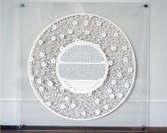 Op de afbeelding: Een witte papieren ketubah met een bloemenontwerp, ingelijst in een helder acryl frame. De ketubah-tekst is in het Hebreeuws en Engels. De tekst luidt: "Dit is de ketubah van het huwelijk van [naam van de bruidegom] zoon van [naam van de vader van de bruidegom] en [naam van de bruid] dochter van [naam van de vader van de bruid]."