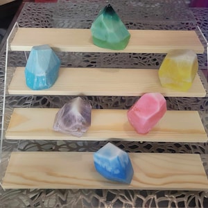 May include: Five colorful geometric shaped soaps displayed on a wooden tiered stand. The soaps are in shades of blue, green, yellow, pink, and purple.