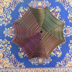 May include: A multi-layered, geometric, star-shaped metal object with a pink and green gradient. The object is made of many thin, concentric rings that create a spiral pattern. The object is on a blue and gold patterned surface.