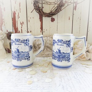 May include: Two white ceramic beer steins with blue floral and windmill designs. Each stein has a handle and the word "Heineken" in a blue decorative frame. The steins are on a white surface with buttons.