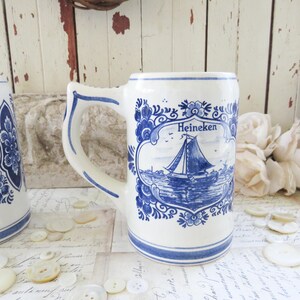 May include: White ceramic beer stein with blue floral and sailboat design. The stein features the word "Heineken" above the sailboat illustration. The handle and rim are accented with blue trim.