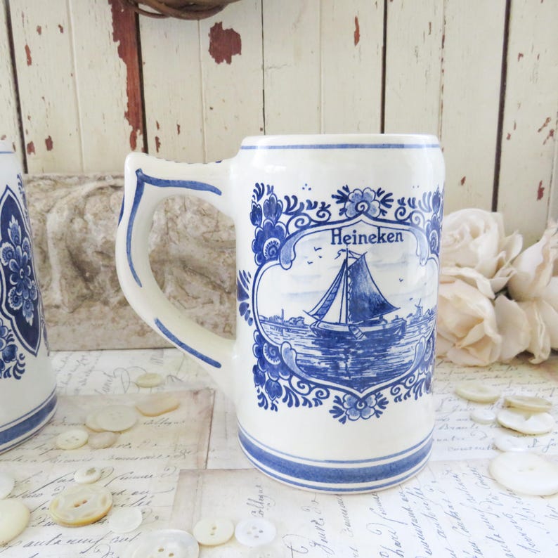 May include: White ceramic beer stein with blue floral and sailboat design. The stein features the word "Heineken" above the sailboat illustration. The handle and rim are accented with blue trim.