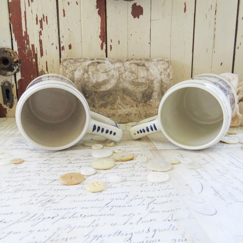 May include: Two ceramic beer steins with handles, featuring a white base with blue and brown decorative bands. The handles have a blue dot design. The steins are resting on a surface with buttons and script.