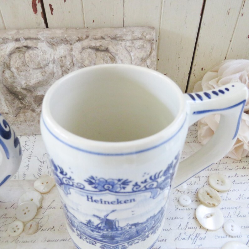 May include: White ceramic mug with blue floral and windmill design, featuring the word "Heineken". The mug has a curved handle and a blue trim. The mug is decorated with a blue and white landscape scene.