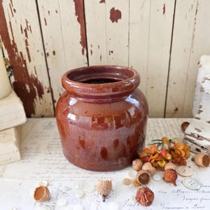May include: A brown glazed ceramic crock with a wide, rounded rim and a cylindrical body. The pot features a smooth, glossy finish with subtle color variations in the clay. It is positioned among scattered white buttons and small acorns.