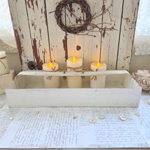 May include: A rustic white wooden tray holds three ivory-colored LED candles with flickering yellow lights. The candles are tied with twine. A small twig wreath hangs above the tray against a distressed white wooden backdrop. The scene evokes a cozy, farmhouse aesthetic.
