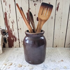 May include: A dark brown cylindrical ceramic crock holding a collection of wooden kitchen utensils. The utensils include a slotted spatula with a charred edge, a wooden spoon, and two thin wooden stirring sticks, all standing upright in the jar.
