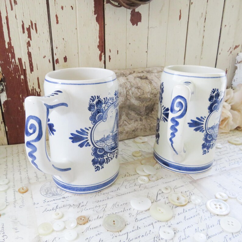 May include: Two white ceramic mugs with blue floral and nautical designs. Each mug has a large handle and a blue rim. The mugs are decorated with a blue and white nautical scene and floral accents.