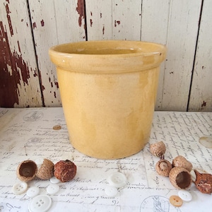 May include: A cylindrical ceramic crock with a smooth, glossy mustard yellow glaze. The vessel features a thick, rounded rim at the top and tapers slightly toward the base. The surface has a uniform, solid color with subtle tonal variations.