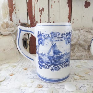 May include: White ceramic beer stein with blue floral and sailboat design. The stein features the word "Heineken" in a decorative frame. The handle and rim are also trimmed in blue. A classic piece for beer enthusiasts.