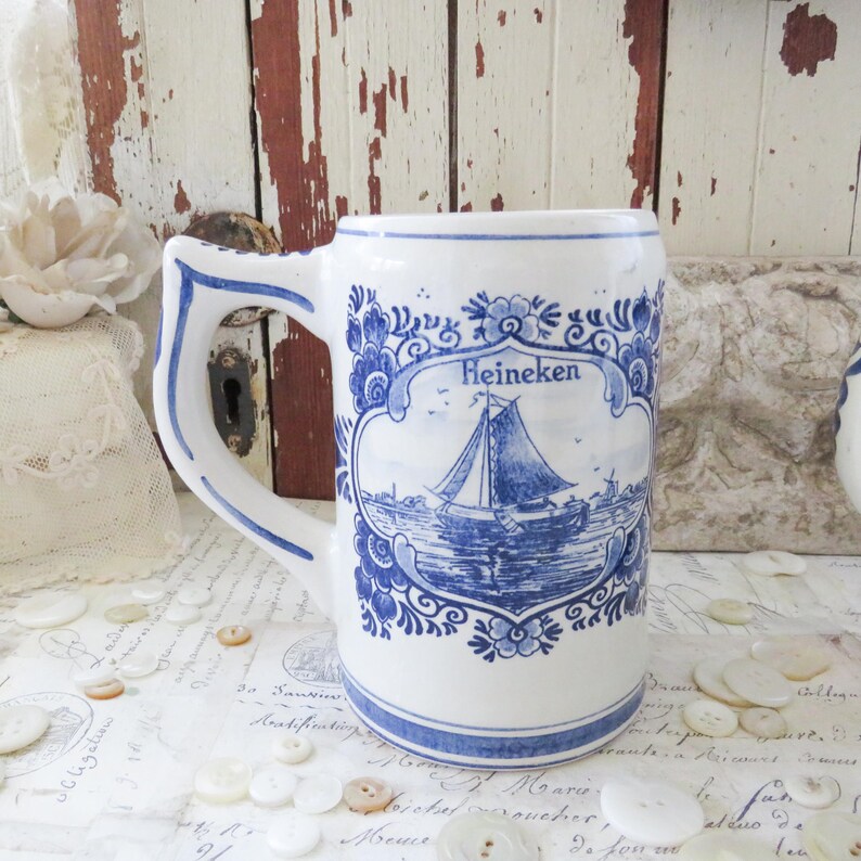 May include: White ceramic beer stein with blue floral and sailboat design. The stein features the word "Heineken" in a decorative frame. The handle and rim are also trimmed in blue. A classic piece for beer enthusiasts.